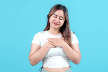 Neşeli bir ifadeyle genç bir kadın, ellerini nazikçe kalbinin üzerine koyuyor, canlı mavi bir arka plana, sıcaklığı ve pozitifliği iletiyor..