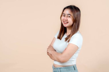 Kollarını kavuşturup gülümseyen kendine güvenen genç bir kadın yumuşak bir bej arka plana karşı gevşek bir tavır sergiliyor. Pozitiflik ve kendine güven yayıyor..