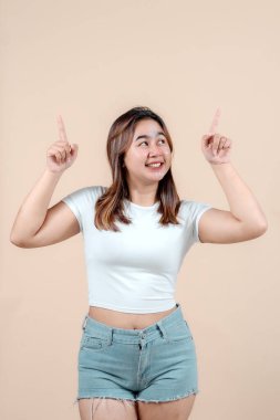 Neşeli genç bir kadın, coşkuyla parmaklarını havaya kaldırarak gülümsüyor, bej bir arka plana yaslanıyor, neşe ve heyecan saçıyor..