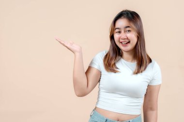 Neşeli genç bir kadın, gülümsüyor ve elini uzatırken şakacı bir şekilde solunu işaret ediyor. Yumuşak bir bej arka plana yaslanmış, neşe ve dostluk saçıyor..