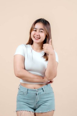 Gülümseyen genç bir kadın olumlu bir jest yapıyor, pozitiflik ve güven yayıyor, bej bej bir arka plana kuruluyor, mutluluk ve onay yayıyor..