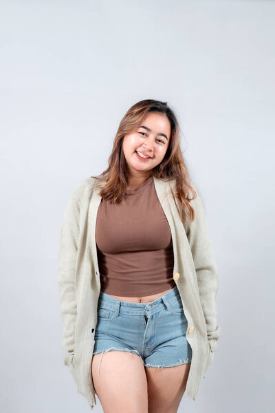 Молодая женщина в уютном свитере и джинсовых шортах уверенно улыбается, демонстрируя непринужденный и стильный вид на нейтральном фоне.