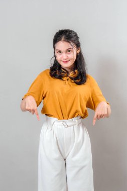 Her iki işaret parmağıyla aşağıya doğru işaret ederken kurnazca gülümseyen kendinden emin genç Asyalı kadın. Dostça bir ifade, reklamcılık, promosyon ve pazarlama için ideal temiz gri bir arkaplan..