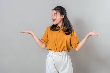 Neşeli, genç Asyalı kadın, yan yan bakarken neşeyle gülümsüyor ve iki elini de oyuncu, anlamlı bir şekilde kaldırıyor, mutluluğu, sürprizi ve dostça bir heyecanı iletiyor..