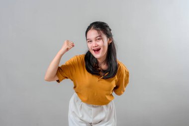 Mutlu, genç Asyalı bir kadın, kendine güven ve başarı göstererek parlak bir gülümsemeyle zafer yumruğu sallıyor. Enerji dolu yüz ifadesi kutlama veya motivasyon temaları için mükemmel.