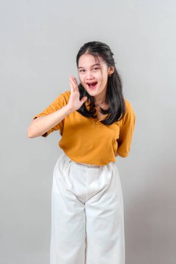 Heyecanlı genç Asyalı kadın, elini ağzının kenarına koyduğu sırada neşeli bir şekilde gülümsüyor, neşeli bir yüz ifadesi sergiliyor, sanki bağırıyor ya da neşeli, canlı bir enerjiyle bağırıyor..