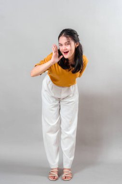 Neşeli ve anlamlı yüzlü genç bir Asyalı kadın elini ağzının kenarına koyuyor, heyecan ve tarafsız bir arkaplan önünde neşeli bir jest gösteriyor..