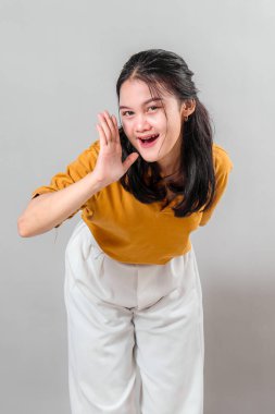 Heyecanlı genç Asyalı kadın, elini ağzının kenarına koyduğu sırada neşeli bir şekilde gülümsüyor, neşeli bir yüz ifadesi sergiliyor, sanki bağırıyor ya da neşeli, canlı bir enerjiyle bağırıyor..
