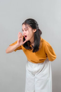 Neşeli ve anlamlı yüzlü genç Asyalı kadın bir eli ağzının kenarında bağırıyor, heyecanla ve neşeyle gülümsüyor, iletişim ya da reklam teması için mükemmel..