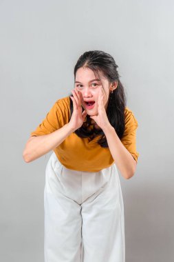 Şaşırmış ve anlamlı yüzlü genç bir Asyalı kadın, elleri ağzının kenarında bağırırken, stüdyo ortamında acil, duyuru veya yüksek sesle iletişim kurarken poz veriyor..