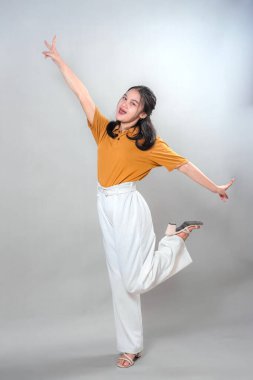 Geniş bir gülümseme ve neşeli bir yüz ifadesiyle, neşeli bir şekilde poz veren genç bir Asyalı kadının tüm vücut portresi, bir bacak kaldırma, kollar yukarı, mutluluk, heyecan ve pozitif enerji yayma.