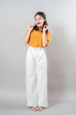 Neşeli, genç Asyalı bir kadının tüm vücut portresi. Parlak, kendinden emin bir gülümsemeyle el hareketi yapıyor. İfade ettiği yüz pozitifliğini, onayını ve dostluğunu gösteriyor..