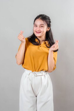 Gülümseyen genç Asyalı kadın iki eliyle parmak kalp hareketi yapıyor. Neşeli yüz ifadesi ve parlak gözleri sevgiyi, pozitifliği ve neşeli arkadaşlığı yansıtıyor..