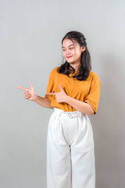 Neşeli, Asyalı genç bir kadın iki eliyle yan tarafı göstererek gülümsüyor. Kendine güveni ve şakacı jesti onu promosyon ya da reklamcılık için ideal yapıyor..