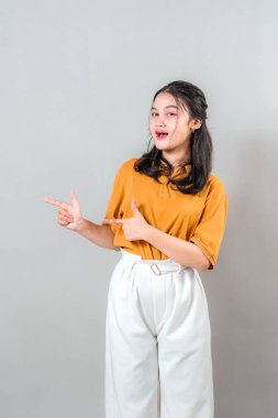 Neşeli, Asyalı genç bir kadın iki eliyle yan tarafı göstererek gülümsüyor. Kendine güveni ve şakacı jesti onu promosyon ya da reklamcılık için ideal yapıyor..