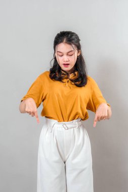 Her iki işaret parmağıyla aşağıya doğru işaret ederken kurnazca gülümseyen kendinden emin genç Asyalı kadın. Dostça bir ifade, reklamcılık, promosyon ve pazarlama için ideal temiz gri bir arkaplan..