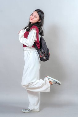 Neşeli genç bir kadın neşeli bir pozla gülümsüyor, bir bacağını kaldırıyor ve ellerini sımsıkı tutuyor, neşe, gençlik ve modern bir görünüşte hakiki mutluluğu ifade ediyor..