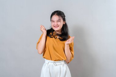Genç Asyalı kadın yoğun heyecan ve neşe ifade ediyor, sıkılmış yumruklarıyla gülümsüyor. Etkileyici yüzü ve enerjik duruşu duygusal pazarlama ve yaratıcı içerik için ideal..