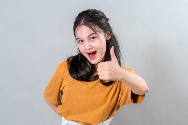 Heyecanlı genç Asyalı kadın, parlak bir ifadeyle başparmak hareketi yapıyor ve ağzı açık. Hevesli yüzü ve enerjik havası reklam ve promosyon için mükemmel..