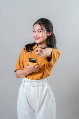 Neşeli genç Asyalı bir kadın kamerayı işaret ederken kendine güvenerek gülümsüyor, akıllı bir telefon tutuyor. Cesaret, onay ya da tanınma gösteren dostane yüz ifadesi.