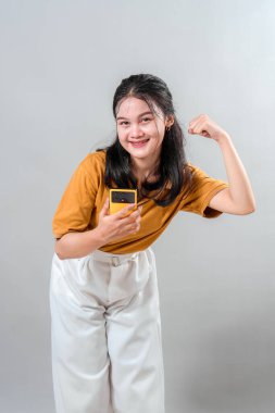 Kolunu esnerken ve akıllı bir telefon tutarken parlak bir şekilde gülümseyen kendine güvenen genç Asyalı kadın. Yüz ifadesi güç, motivasyon ve kendine güven yayıyor..