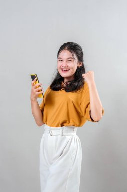 Heyecanlı genç Asyalı kadın akıllı telefonuna muzaffer bir yüz ifadesiyle bakıyor. Gülümser ve yumruğunu sıkar, neşe, başarı ve duygusal zafer gösterir..