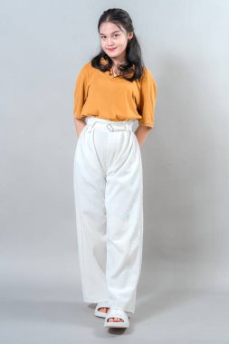 Kendine güvenen ve çekici bir yüz ifadesiyle gülümseyen genç Asyalı kadın elleri ceplerinde gelişigüzel duruyor. Stüdyo portresi doğal sıcaklığı ve arkadaş canlısı kişiliğini gösteriyor..