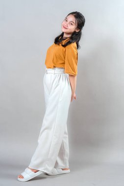 Genç Asyalı kadının portresi vücudunu eğerek gülümserken nazik ve kendinden emin bir ifadeyle. Tarafsız arkaplanlı stüdyo çekimi, sıcaklık ve pozitif enerji aktarımı.