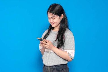 Uzun siyah saçlı Endonezyalı genç bir kadın, günlük gri bir tişört giyiyor, parlak mavi bir arka planda akıllı telefonu tutarken gülümsüyor. Kendinden emin ve rahat bir duruş..