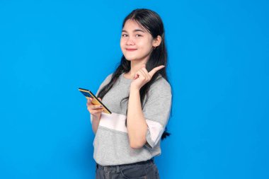 Endonezyalı, uzun siyah saçlı, gri tişörtlü, elinde akıllı bir telefonla gülümseyen ve parlak mavi bir arkaplanı işaret eden genç bir kadın. Neşeli ve kendinden emin..