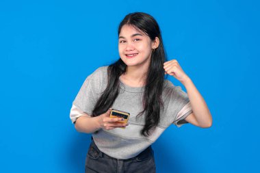 Endonezyalı genç bir kadın, uzun siyah saçlı, gri tişörtlü, akıllı telefonu tutarken gülümseyen ve heyecan içinde yumruğunu kaldıran parlak mavi bir arka planda..