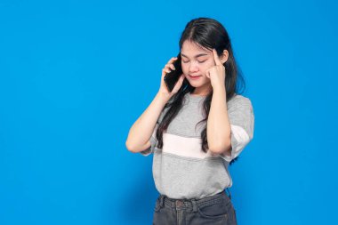 Uzun siyah saçlı Endonezyalı genç bir kadın, gri bir tişört giyiyor, telefonda düşünceli ve duygusal bir ifadeyle konuşuyor, şakaklarına dokunuyor, mavi bir arka planda..