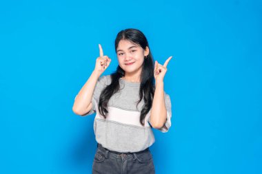 Uzun siyah saçlı, gri bir süveter giyen Asyalı genç bir kadın. Güvenle gülümseyen ve her iki işaret parmağını yukarı doğru gösteren bir kadın..