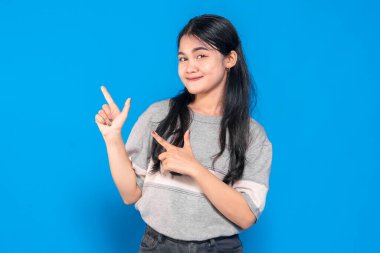 Uzun siyah saçlı, gri tişörtlü bir Asyalı genç kadın, iki eliyle mavi arka planda işaret ederken neşeli bir şekilde gülümsüyor..