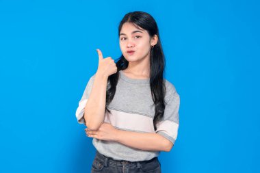 Neşeli bir gülümsemesi olan genç bir Asyalı kadın, mavi bir arkaplana karşı dururken baş parmak işareti yapıyor. Uzun siyah saçları var ve günlük gri tişört ve siyah kot pantolon giyiyor..