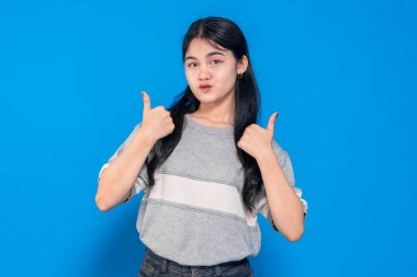Neşeli bir gülümsemesi olan genç bir Asyalı kadın, mavi bir arkaplana karşı dururken baş parmak işareti yapıyor. Uzun siyah saçları var ve günlük gri tişört ve siyah kot pantolon giyiyor..