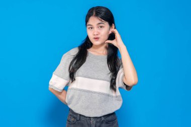 Genç Asyalı kadın parmaklarıyla kalp şekli oluştururken neşeli bir surat ifadesi takınıyor. Uzun siyah saçları var, günlük gri bir tişört giyiyor ve mavi arka planda duruyor..