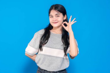 Genç Asyalı kadın gülümsüyor ve iyi bir el hareketi yapıyor. Uzun siyah saçları var, günlük gri bir tişört giyiyor ve mavi bir arka plana karşı duruyor, pozitifliğini gösteriyor..