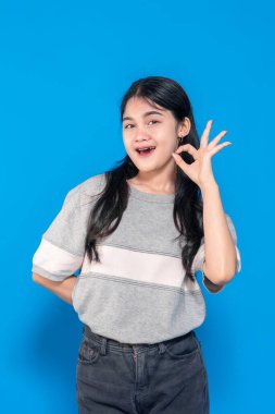 Genç Asyalı kadın gülümsüyor ve iyi bir el hareketi yapıyor. Uzun siyah saçları var, günlük gri bir tişört giyiyor ve mavi bir arka plana karşı duruyor, pozitifliğini gösteriyor..