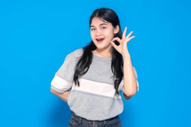 Genç Asyalı kadın gülümsüyor ve iyi bir el hareketi yapıyor. Uzun siyah saçları var, günlük gri bir tişört giyiyor ve mavi bir arka plana karşı duruyor, pozitifliğini gösteriyor..