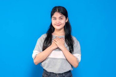 Uzun siyah saçlı genç Asyalı kadın ellerini göğsüne koyarken nazikçe gülümsüyor. Gri bir süveter giyiyor ve mavi bir arka plana karşı duruyor. Minnettarlığını ifade ediyor..