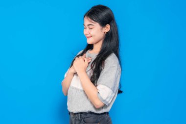 Uzun siyah saçlı genç Asyalı kadın nazikçe gülümsüyor ve elleri göğsünde, minnettarlığını ve huzurunu ifade ediyor. Mavi arka planda sıradan bir kıyafet giyiyor..