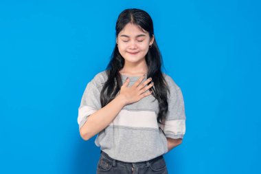 Sıcak bir gülümseme ile genç bir Asyalı kadın, bir elini göğsüne koyuyor, minnettarlığını ve samimiyetini gösteriyor. Sıradan bir kıyafet giyiyor ve parlak mavi bir arka plana karşı duruyor..