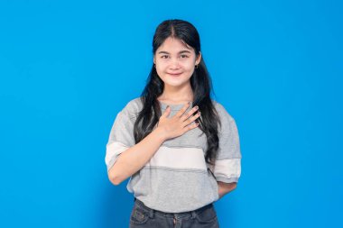 Sıcak bir gülümseme ile genç bir Asyalı kadın, bir elini göğsüne koyuyor, minnettarlığını ve samimiyetini gösteriyor. Sıradan bir kıyafet giyiyor ve parlak mavi bir arka plana karşı duruyor..