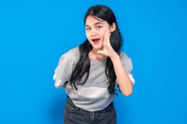 Genç Asyalı kadın neşeli bir ifadeyle neşe içinde bağırıyor. Elini ağzının yanında tutuyor, sanki bağırıyor gibi, parlak mavi bir arka plana karşı heyecanlı ve anlamlı görünüyor..