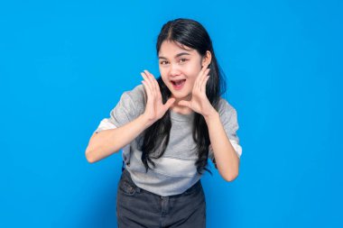 Heyecanlı bir suratı olan genç bir Asyalı kadın, elleri yüzünün yanında bağırıyor ya da bir şey söylüyor gibi. Parlak mavi bir arka planda neşeli ve enerjik görünüyor..