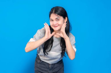 Heyecanlı bir suratı olan genç bir Asyalı kadın, elleri yüzünün yanında bağırıyor ya da bir şey söylüyor gibi. Parlak mavi bir arka planda neşeli ve enerjik görünüyor..