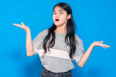 Kafası karışmış genç bir Asyalı kadın, sorgulama jesti için iki elini de kaldırıyor. O, parlak mavi bir arka plan karşısında duran, belirsiz ya da şaşırmış görünüyor.