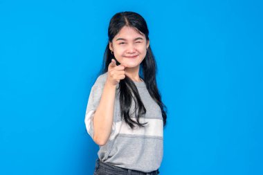 Genç Asyalı kadın neşeli bir şekilde gülümsüyor, kendinden emin bir şekilde kameraya bakıyor. Neşesini ve dostluğunu ifade eder, parlak mavi bir arka plana karşı durarak..