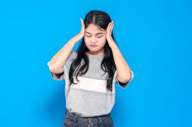 Stresli görünen genç bir Asyalı kadın, iki eli kapalı, gözleri kapalı ve dudakları sıkılmış bir şekilde başını tutuyor. Hüsran ve endişeyi canlı, mavi bir arka plana karşı ifade eder..